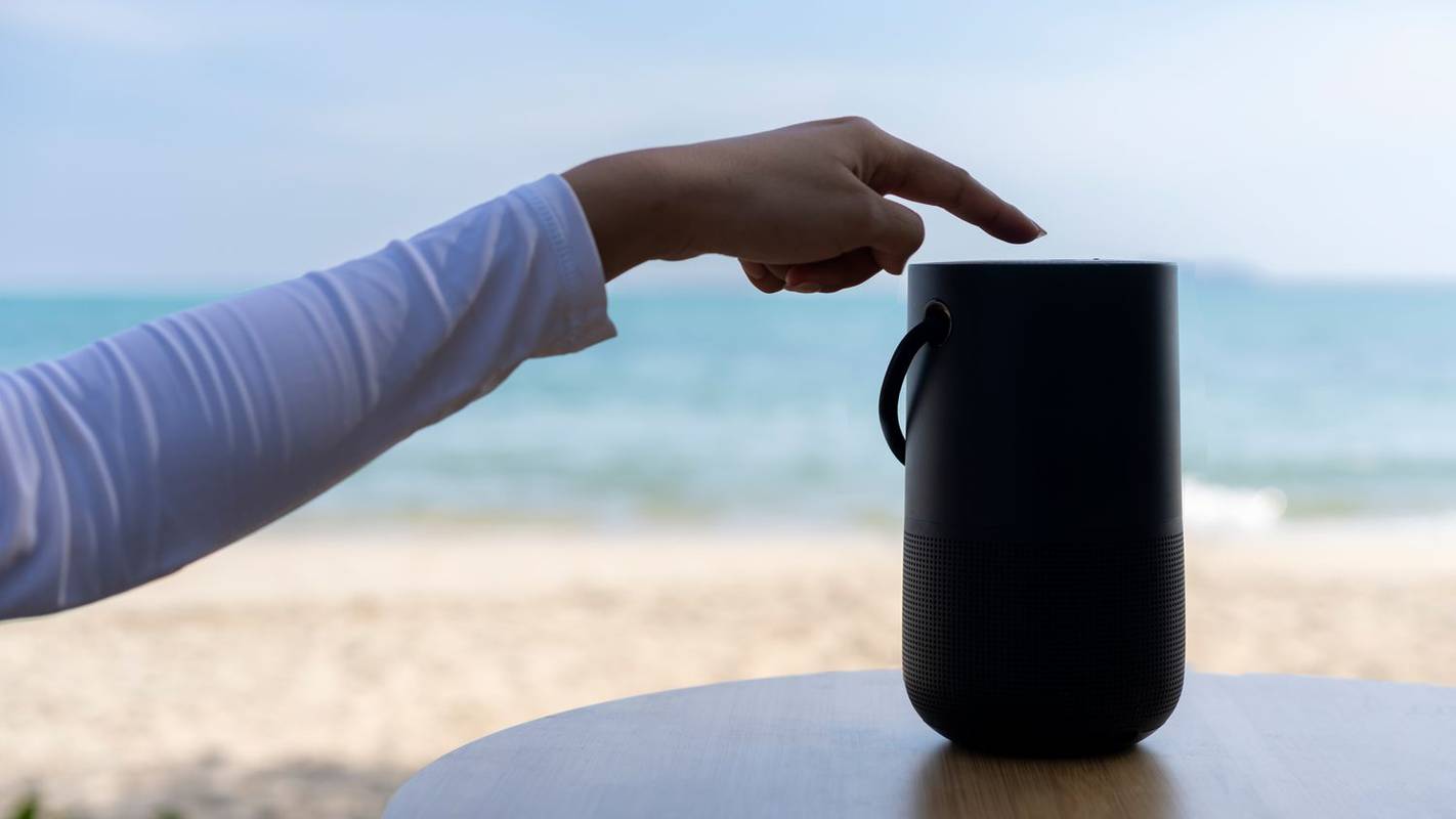 Sebuah tangan menyalakan speaker Bluetooth dengan latar belakang pantai.